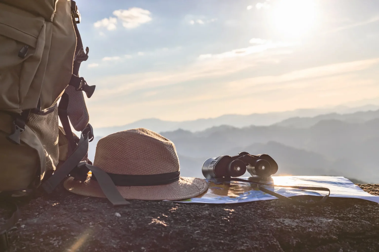 Un sac à dos, un chapeau de randonnée et des jumelles posées sur un rocher qui surplombe un paysage dégagé avec un rayon de soleil qui symbolise l'ouverture vers les possibles et l'exploration nécessaire pour y arriver