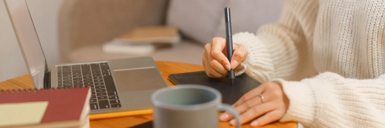 des mains de femme qui dessin avec devant elle le clavier de son ordinateur et une tasse en 1er plan