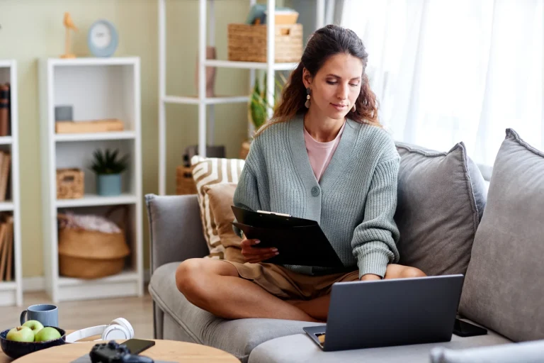 Une femme est assise sur son cananpé chez elle. Elle a dans les mains des documents et consulte son ordinateur portable posé sur son canapé. Derière elle le décors est chaleureux et cocooning.
