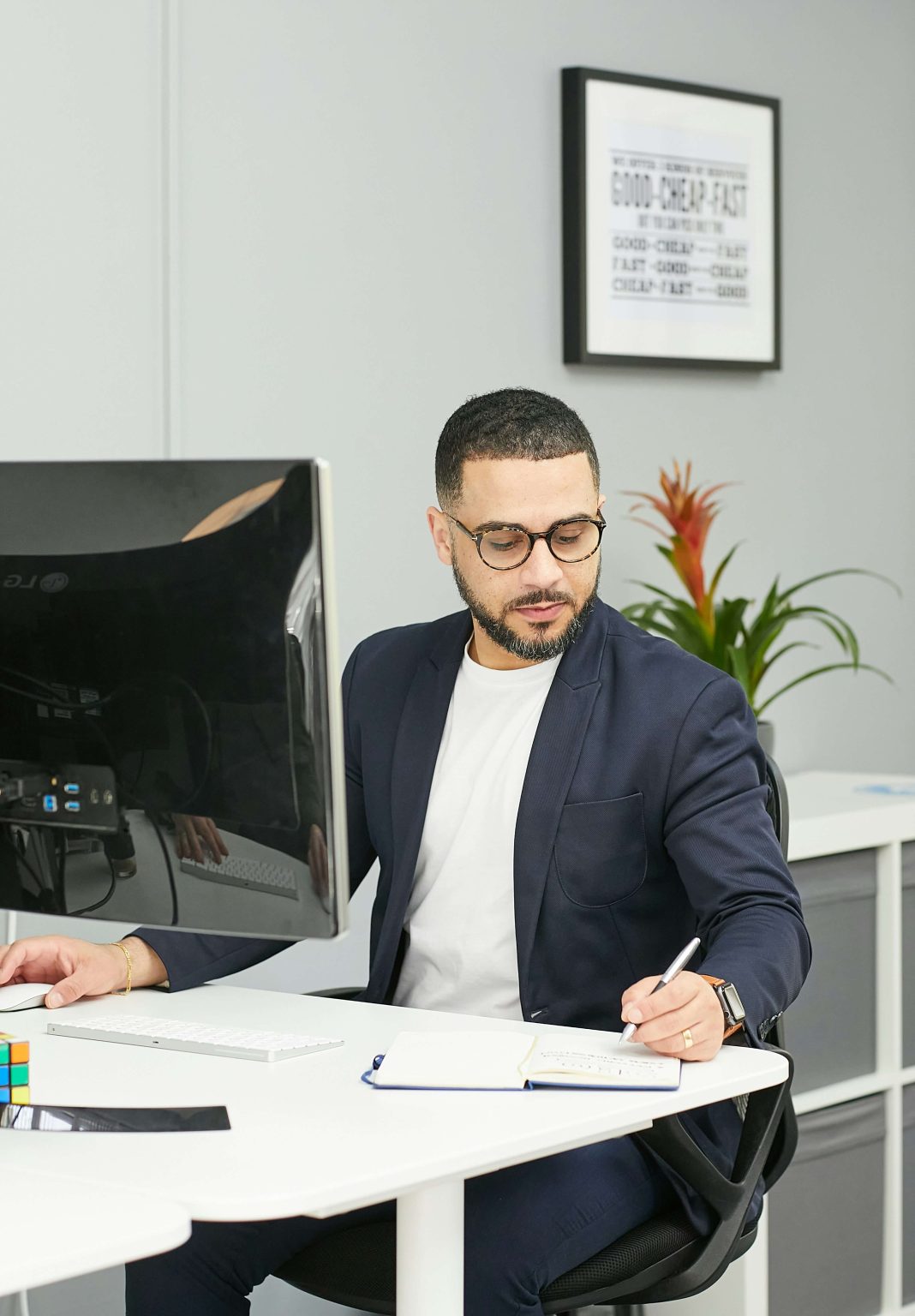 un homme de 40 ans environ est assis devant son ordinateur il prend des notes sur un carnet posé à côté de lui. Il semble concentré sur son travail.